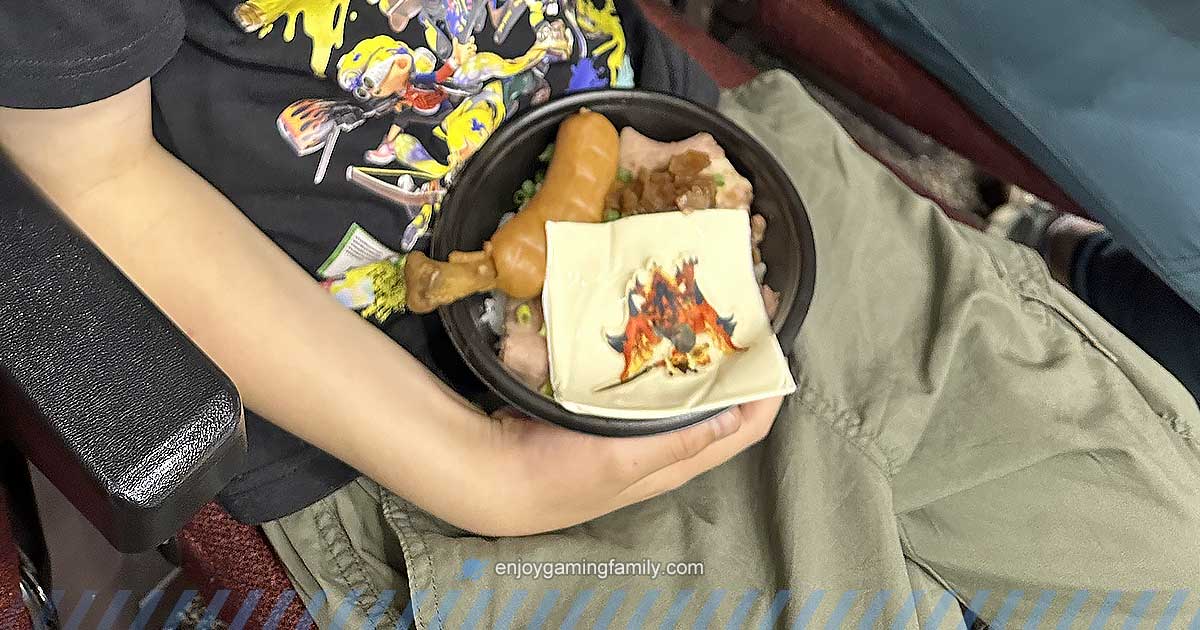 東京ゲームショウ2024のファミリーゲームパークで、子どもがローストビーフ丼を膝の上に乗せて座席で食べている様子。モンハンのリオレウスがプリントされたチーズがトッピングされている。