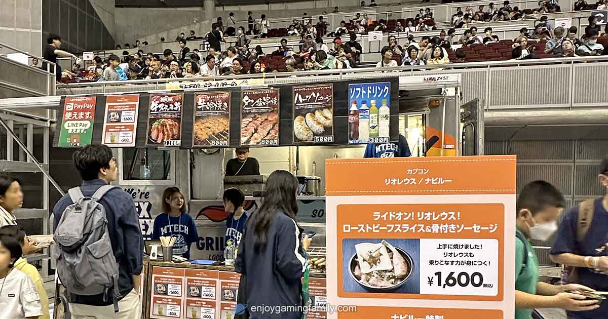 東京ゲームショウ2024のファミリーゲームパーク内にあるキッチンカー。カプコンのコラボメニュー『ローストビーフ＆骨付きソーセージ丼（1600円）』の看板が目立ち、後方には観客席で食事中の親子連れの姿。