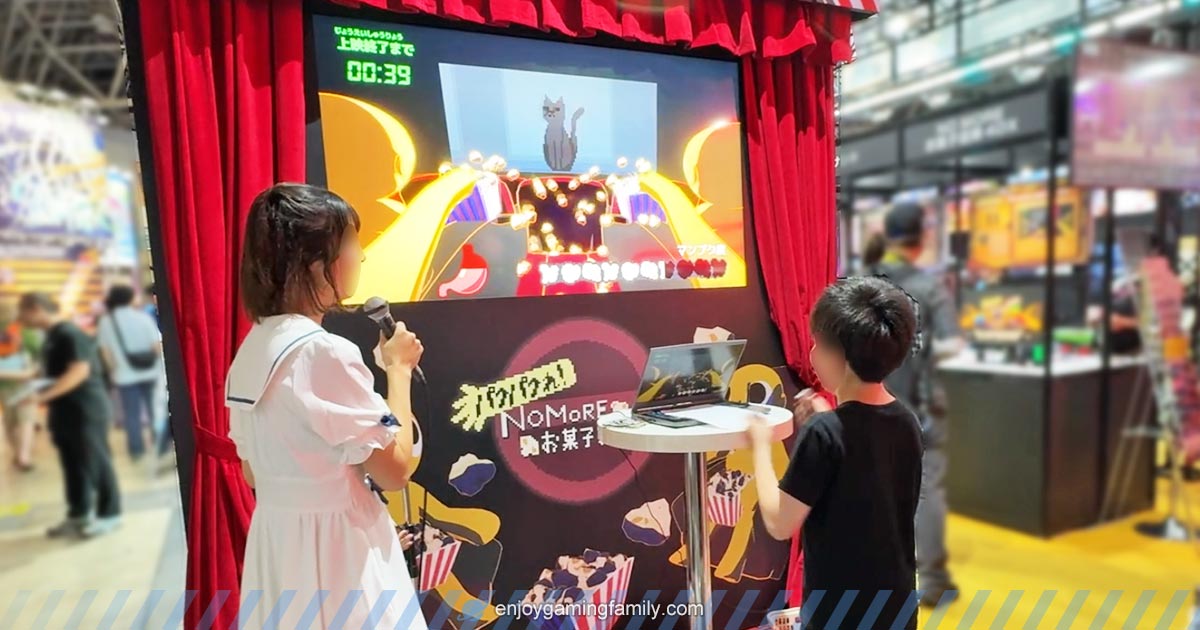 No More お菓子泥棒 4DX』の体験ブースで、子どもがゲームをプレイし、司会の女性がマイクで盛り上げている様子。
