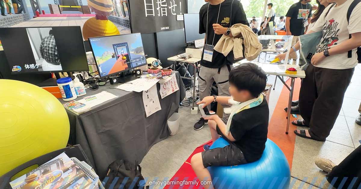 BB Adventureを試遊中の子どもが、バランスボールに乗って体を動かしながらプレイしている様子。モニターにはゲーム画面、周囲には見学者やスタッフがいる。