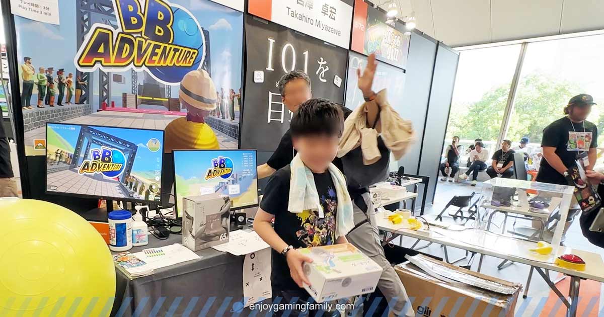BB Adventureを体験したあと、景品を手にして喜ぶ子どもとスタッフ。ブースにはゲームロゴと大型モニターがあり、賑やかな雰囲気。